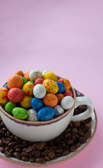 Composition with small chocolate easter eggs in cup surrounded by coffee roasted beans. Easter coffee concept. Mug full of colorful eggs on a plate with coffee beans on a pink background.