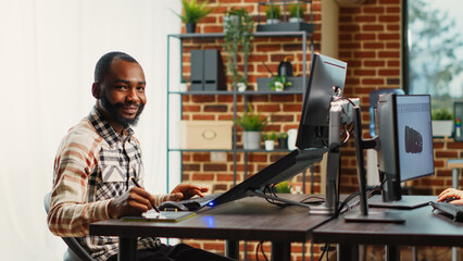 Happy content creator developing software interface on touchscreen display, using tablet and stylus at desk. African american man working on infographics production in agency studio.