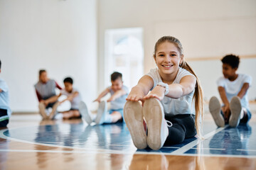 Happy schoolgirl stretching during PE class and looking ta camera. - obrazy, fototapety, plakaty