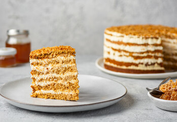Piece of biscuit cake Medovik. Layered caramel honey cake with whipped cream and sour cream. Honeycomb and jars of honey on concrete background.