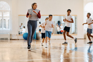 Happy PE teacher and group of kids running during exercise class at school gym.