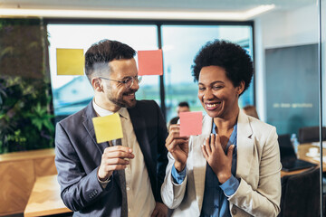 Young woman of a small company putting an adhesive note on glass in office during team meeting formulating business strategies.