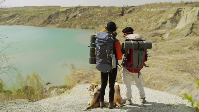 Full back shot of two dark-haired tourists with rucksacks standing on rocky coast of lake, two small dogs beside, looking at camera photos together, head to head. Windy weather, day
