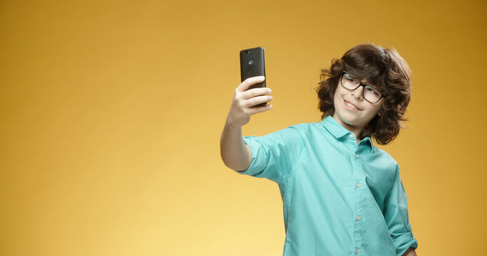 Cute little boy using his mobile phone to take pictures or selfies and positively smiling, isolated on yellow background - always online, emotions concept close up 