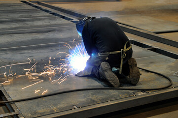 Welding process. A welder on his knees welds metal structures in a factory.