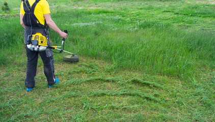 A male gardener mows the green grass of the lawn in the backyard with a gasoline mower. Trimmer for the care of a garden plot