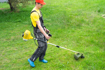A male gardener mows the green grass of the lawn in the backyard with a gasoline mower. Trimmer for...