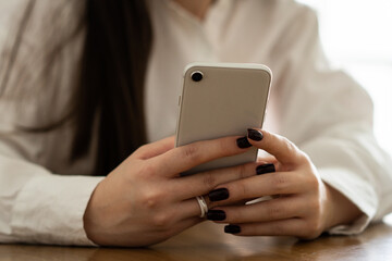 Cropped view of a woman in a white shirt who holds a white smartphone in her hands. A brunette woman sitting at a table uses a smartphone.