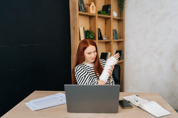 Front view of young woman with broken hand wrapped in white gypsum bandage having online video...