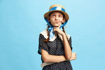 Pretty young girl wearing old style clothes holding finger near chin and looking up with no sure facial expression over blue background. Doubt, uncertainty, pensive