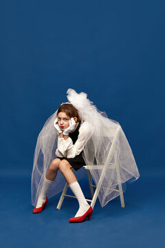 Portrait of young girl wearing brides veil sitting on chair with sad facial expression over blue background. Little sad bride