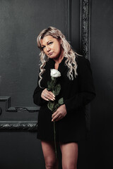Portrait of pensive middle aged 45 years old woman in black with white rose posing in dark room, looking at camera. Thoughtful sad alone mature lady standing at black wall. Emotion concept. Copy space