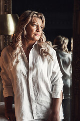 Pensive middle aged 45 years old woman standing in dark living room with mirror, thinking looking away. Thoughtful alone mature lady in white clothes posing. Loneliness concept. Copy ad text space
