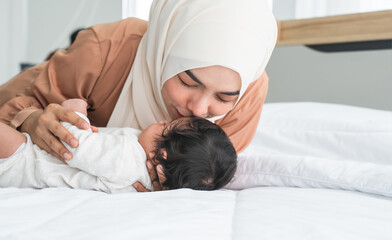 Asian young Muslim mother wearing hijab kissing her newborn baby 1 month old on forehead while cute infant sleeping on bed in bedroom at home. Muslim family, child care and mother's day concept