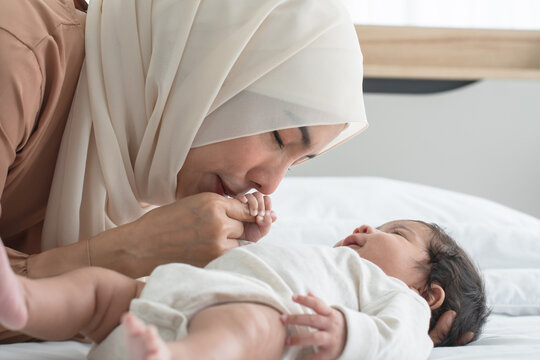 Asian young Muslim mother wearing hijab kissing hand of her newborn baby 1 month old while cute infant grabbing mom finger, sleeping on bed in bedroom. Muslim family, child care, mother's day concept