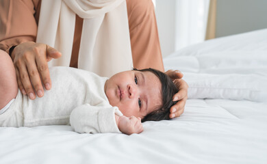 Obraz na płótnie Canvas Portrait of adorable Asian Muslim newborn baby 1 month old lying on bed, looking at camera with innocent face. Mother hands is lull her little daughter infant by tender touching head and body