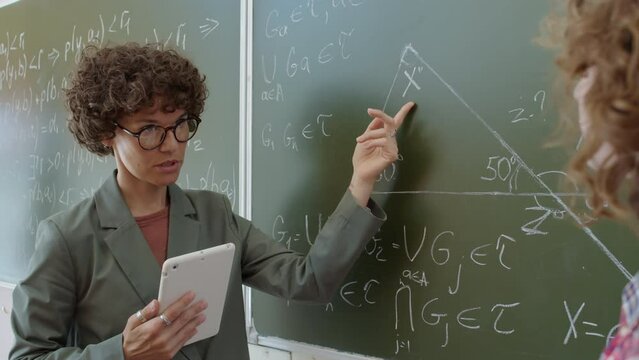 Female Math teacher standing in front of her student explaining how to solve geometry problem written on blackboard
