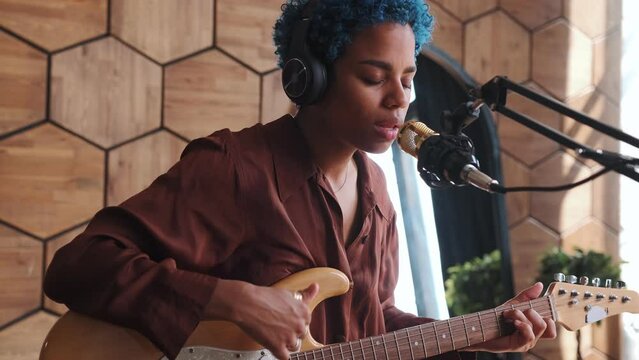 Young creative beautiful African American woman singer with blue hair plays guitar and performs songs in style of POP or country music recording new album sits in recording studio with microphone