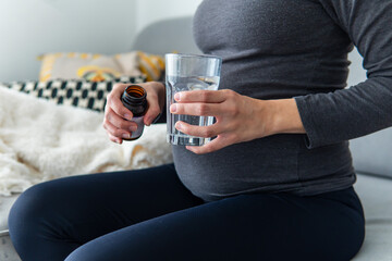 Pregnant woman taking vitamins pills and minerals from a jar with a glass of water while sitting comfortably on the sofa. Pregnancy, healthcare, and supplements concept. Woman taking medicine at home.