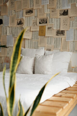 Fototapeta na wymiar Comfy white bed, wall covered with pages from books and House plant. Morning in bright boho chic style bedroom. Scandinavian interior design