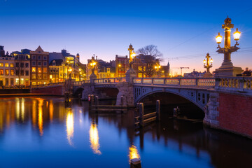 Naklejka na ściany i meble Blue bridge, Amsterdam, Netherlands. Blauwbrug. Evening cityscape. Blue sky and city lights. Dutch canals. Reflections on the surface of the water. Photography for design and wallpaper.