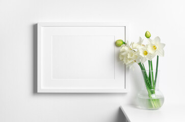 Blank photo frame mockup in white interior with narcissus flowers in vase