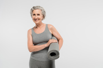 cheerful senior woman in sportswear holding fitness mat and smiling isolated on grey.