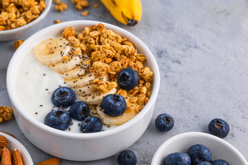 Bowl of oat granola with yogurt, banana, blueberries, chia seeds and nuts on gray background for healthy breakfast
