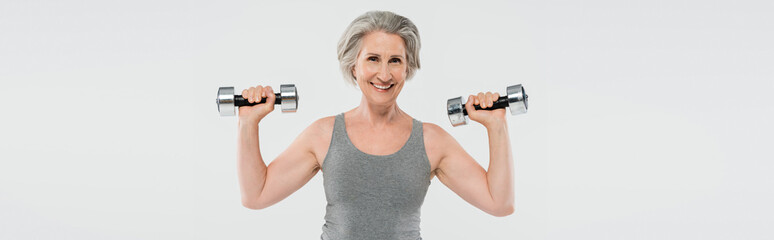 overjoyed senior woman with grey hair exercising with dumbbells isolated on grey, banner.