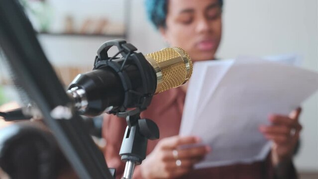 Close up professional recording microphone and woman with sheets of paper reading latest current news to notify listeners of important events or breaking news sits in studio or home office