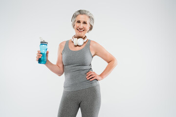 happy senior woman with wireless headphones holding sports bottle and standing with hand on hip isolated on grey.