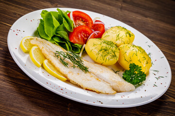 Fish dish - fried cod with boiled potatoes and fresh vegetables on wooden table
