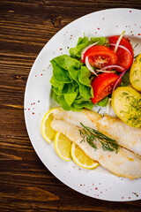 Fish dish - fried cod with boiled potatoes and fresh vegetables on wooden table
