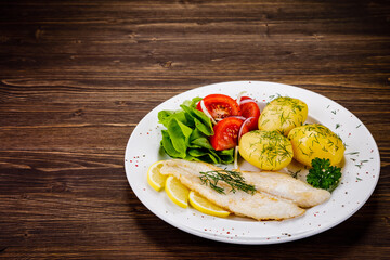 Fish dish - fried cod with boiled potatoes and fresh vegetables on wooden table
