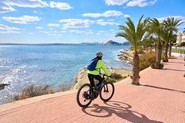 Obraz premium nice senior woman cycling on a cool spring day with her electric bicycle at the waterfront of Alicante, Costa Blanca, Spain