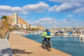 nice senior woman cycling on a cool spring day with her electric bicycle at the waterfront of Alicante, Costa Blanca, Spain - obrazy, fototapety, plakaty