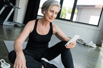 senior sportswoman using mobile phone and sitting on fitness mat in gym.