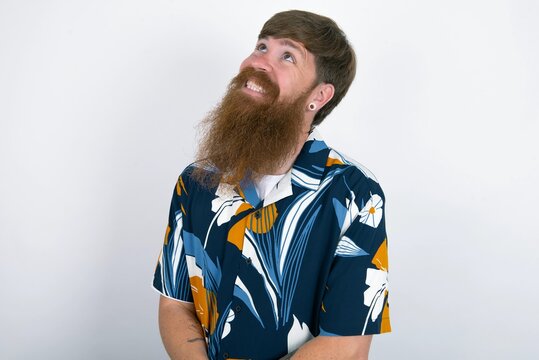 Portrait of mysterious red haired man wearing printed shirt over white studio background looking up with enigmatic smile. Advertisement concept.