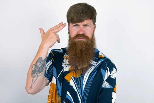 Unhappy red haired man wearing printed shirt over white studio background imitates gun shoot makes suicide gesture keeps two fingers on temples.