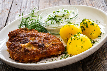 Breaded fried pork chop with fresh cucumber in cream and potatoes on wooden table 