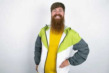 Studio shot of cheerful red haired man wearing sportswear standing over white studio background keeps hand on hip, smiles broadly.