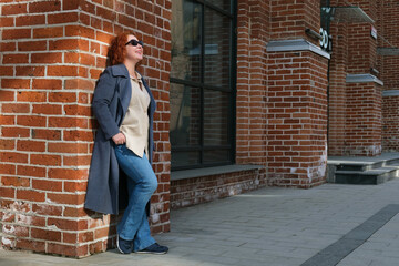 Fototapeta na wymiar Attractive red-haired woman in a blue coat, jeans stands outside a brick building. Woman in sunglasses looks up and smiles. Portrait of a woman in full growth