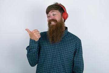 red haired man wearing plaid shirt over white studio background listens audio track via wireless headphones points thumb away advertises copy space