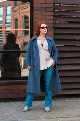 Attractive red-haired woman in a blue coat, jeans and a loose shirt stands outside a brick building with mirrored facade. A woman in sunglasses looks away. Vertical portrait of a woman in full growth