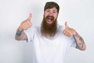 red haired man wearing white T-shirt over white studio background approving doing positive gesture with hand, thumbs up smiling and happy for success. Winner gesture.
