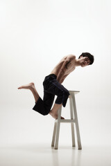 Image of handsome young man posing shirtless in black trousers, jumping on high chair against white studio background. Concept of male body aesthetics, style, fashion, health, men's beauty