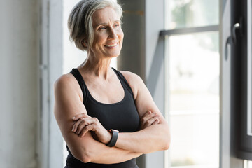 happy senior sportswoman in tank top smiling while standing with crossed arms in gym.