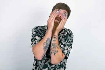 red haired man wearing printed shirt over white studio background keeps hands on cheeks has bored displeased expression. Stressed hopeless model