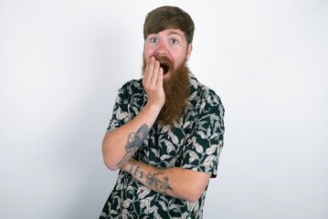 red haired man wearing printed shirt over white studio background covers mouth and looks with wonder at camera, cannot believe unexpected rumors.