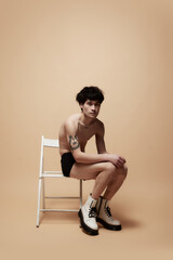 Shy look. Young asian guy in underwear and stylish boots, posing shirtless on chair against light brown studio background. Concept of male body aesthetics, style, fashion, health, men's beauty